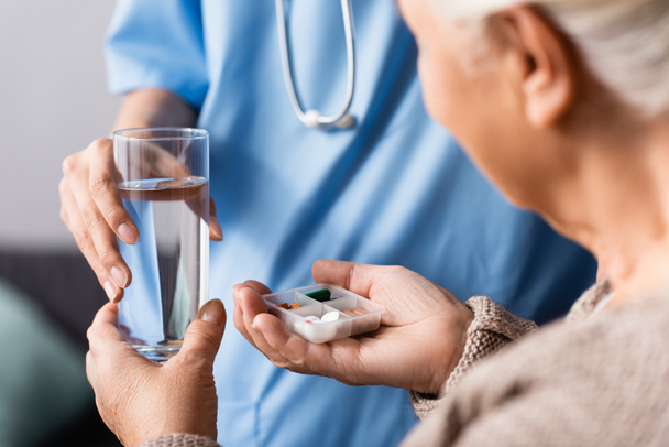 visão parcial da enfermeira dando pílulas e copo de água para a mulher idosa, foco seletivo - Foto, Imagem