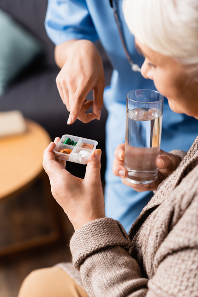 enfermera señalando píldoras en la mano de la mujer mayor con vaso de agua, fondo borroso - Foto, Imagen