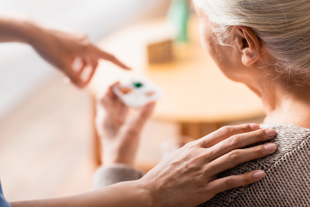 partial view of nurse touching shoulder of aged woman and pointing with finger at pills on blurred background - Photo, Image