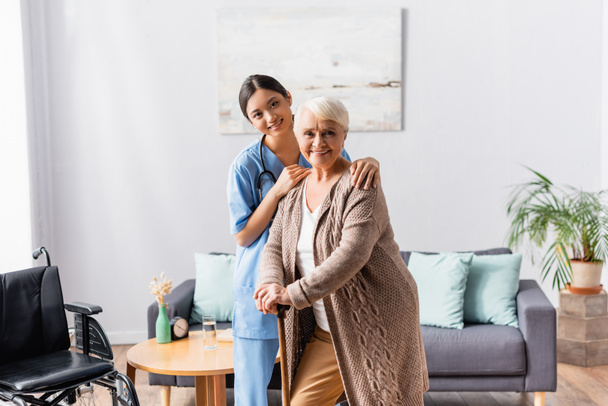 smiling asian nurse and elderly woman with walking stick looking at camera in nursing home - Photo, Image