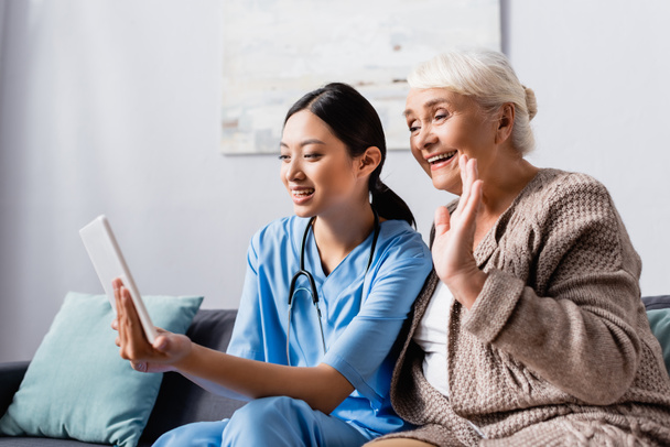 усміхнена азіатська медсестра тримає цифровий планшет біля щасливої старшої жінки махає рукою під час відео розмови
 - Фото, зображення