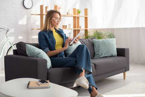 Young blonde woman in eyeglasses using digital tablet while sitting on couch at home on blurred background - Foto, Imagem