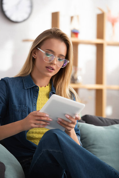 Blonde woman in eyeglasses looking at digital tablet while sitting on couch on blurred background - Foto, Imagem