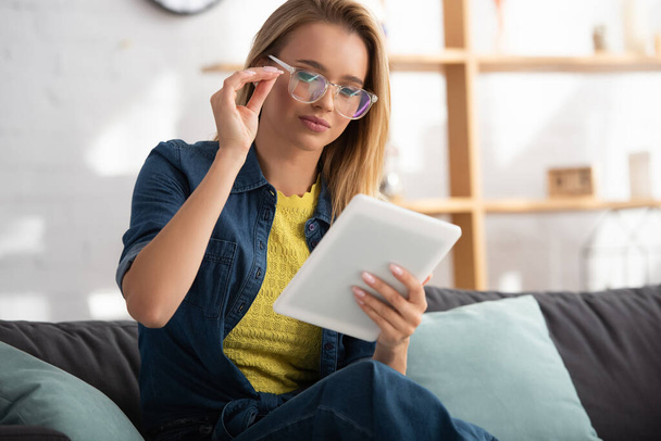 Young blonde woman touching eyeglasses while looking at digital tablet at home on blurred background - Foto, Imagem