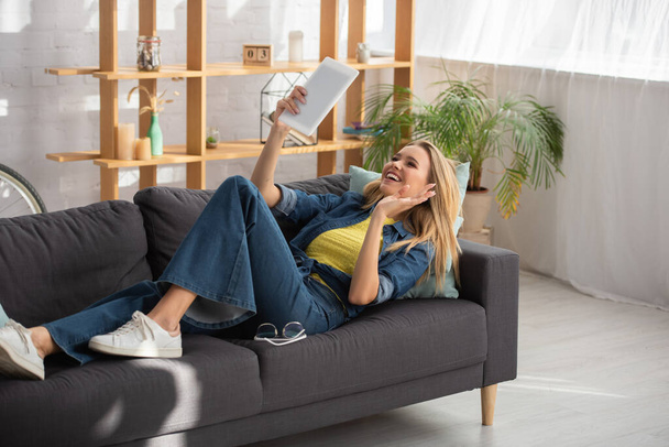 Femme blonde souriante avec main agitant regardant la tablette numérique tout en étant allongé sur le canapé à la maison - Photo, image