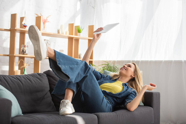 Молода блондинка дивиться на цифровий планшет, лежачи на дивані вдома на розмитому фоні
 - Фото, зображення
