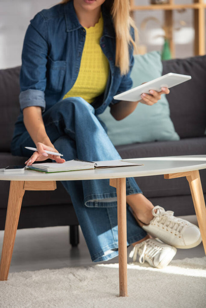 Vista recortada de una mujer joven con lápiz y tableta digital hojeando el cuaderno en casa sobre un fondo borroso - Foto, imagen