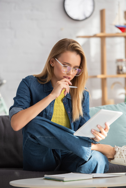 Thoughtful blonde woman with pen looking at digital tablet while sitting on couch on blurred background - Foto, afbeelding