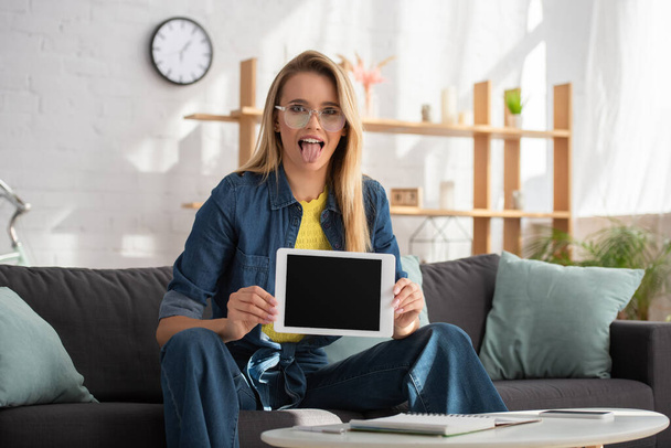 Young blonde woman with sticking out tongue looking at camera while showing digital tablet at home on blurred background - Foto, Imagem