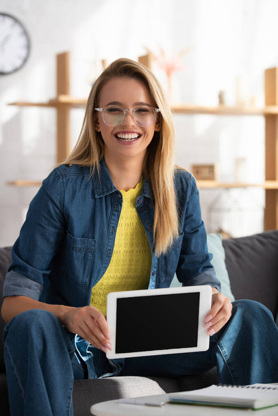 Cheerful young blonde woman looking at camera while showing digital tablet at home on blurred background - Fotoğraf, Görsel