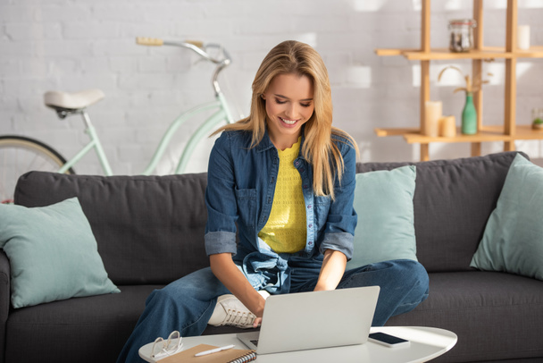 Mulher loira alegre usando laptop enquanto sentado no sofá em casa no fundo borrado - Foto, Imagem