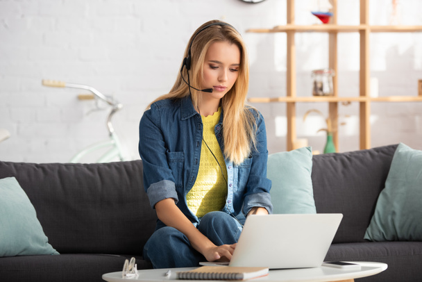 Young blonde woman in headset typing on laptop while sitting on couch on blurred background - Fotó, kép