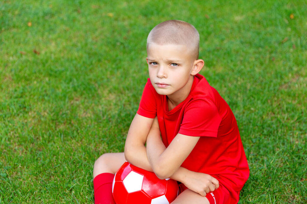 Een jongen in een rood voetbaluniform met een bal in zijn handen zit op het voetbalveld. Kijk naar de camera. Serieuze blik - Foto, afbeelding