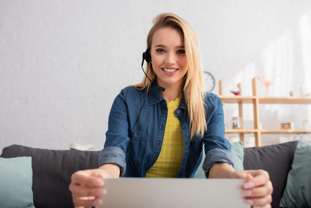 glimlachende blonde vrouw in headset kijken naar camera terwijl het houden van laptop op wazig achtergrond - Foto, afbeelding