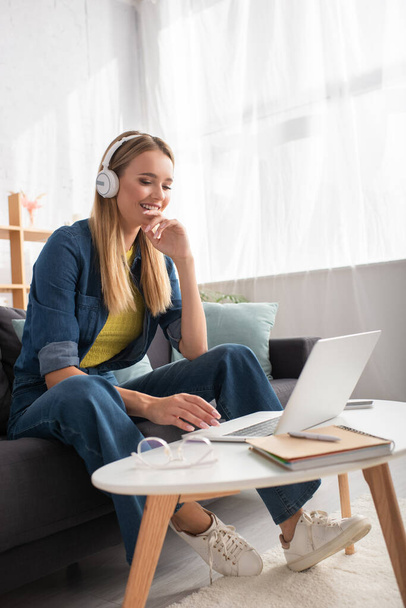 Mujer joven feliz en los auriculares mirando a la computadora portátil mientras está sentado en el sofá cerca de la mesa de café sobre fondo borroso - Foto, Imagen