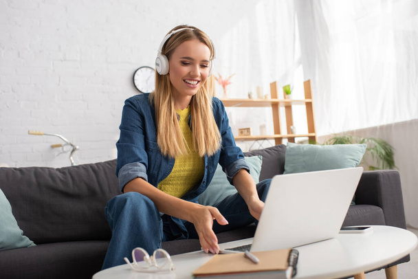 Fröhliche blonde Frau mit Kopfhörer blickt auf Laptop, während sie auf Couch neben Couchtisch auf verschwommenem Hintergrund sitzt - Foto, Bild