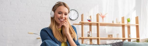 Cheerful young blonde woman in headphones at home on blurred background, banner - Photo, Image