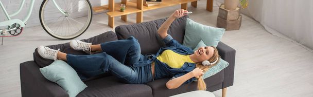 High angle view of happy blonde woman with smartphone laughing while lying on couch at home, banner - Фото, изображение