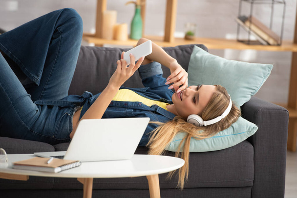 Smiling blonde woman in headphones looking at smartphone while lying on couch near coffee table on blurred background - Fotó, kép