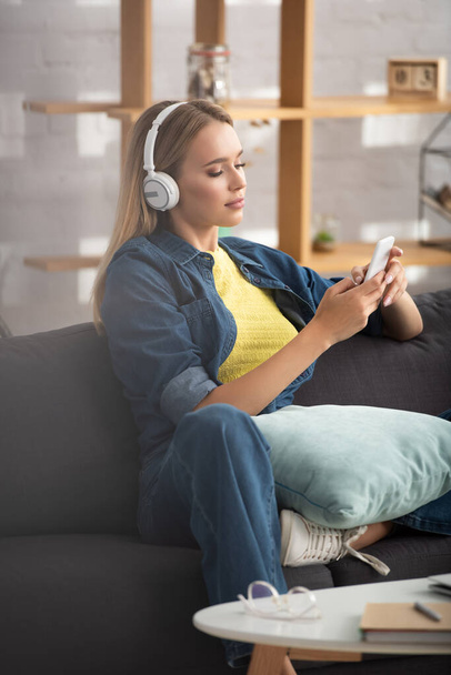 Young blonde woman in headphones texting on smartphone while sitting on couch on blurred background - Foto, imagen