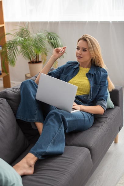 Full length of young woman looking away while using laptop on couch at home - Foto, immagini
