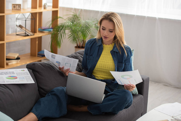 jong blond vrouw met papieren kijken naar laptop terwijl zitten op bank thuis op wazig achtergrond - Foto, afbeelding