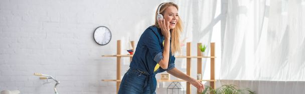 Happy cheerful blonde woman in headphones laughing on blurred background at home, banner - Foto, Bild