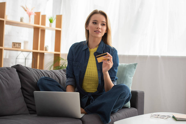 Young blonde woman with credit card looking away while sitting near laptop on couch on blurred background - Fotografie, Obrázek
