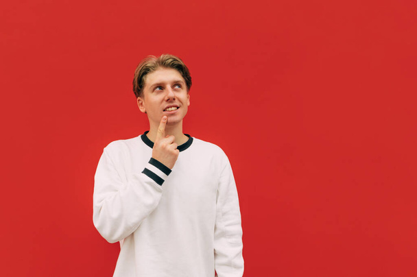 Positive pensive guy in a white sweatshirt stands on a background of a red wall with a finger attached to his lips and looks away with a smile on his face. - Photo, Image