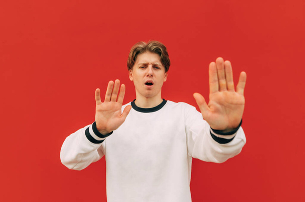 Young man stands on a background of a red wall, looks at the camera with a surprised face and shows his palms gesture Stop. Guy shows his palms to the camera and says stop. Isolated. - Photo, Image