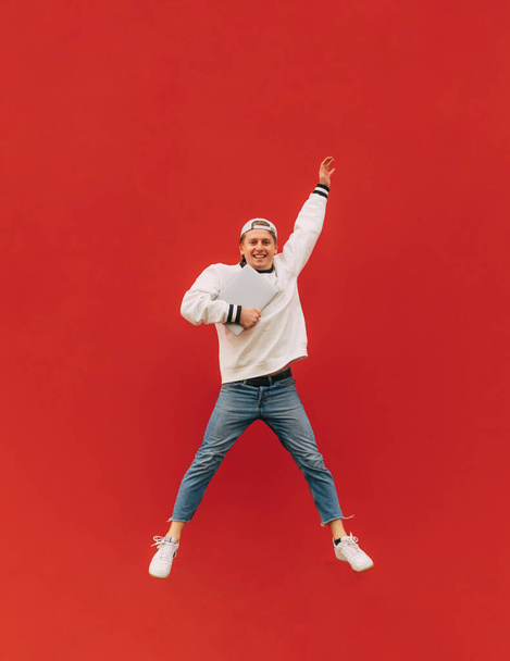 Young man in stylish clothes jumps with a laptop in his hands on a red background, looks at the camera and smiles. Student guy in jump isolated. Vertical. - Photo, Image