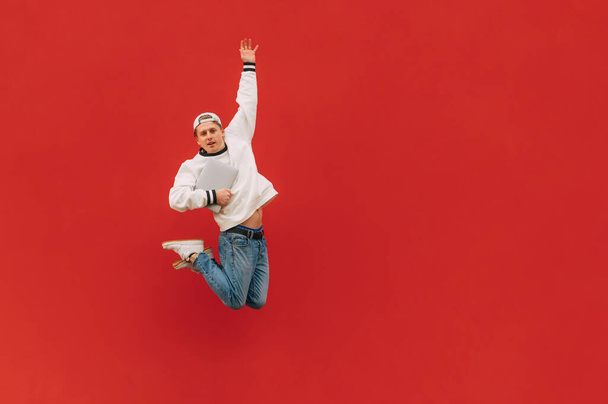 Позитивный молодой человек в уличной одежде с ноутбуком в руках прыгает на фоне красной стены, глядя в камеру. Парень левитирует с ноутбуком на красном фоне - Фото, изображение