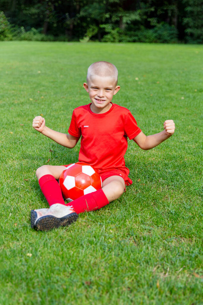 Poika punaisessa jalkapallo-univormussa istuu jalkapallokenttä, iloisesti nosti kätensä - Valokuva, kuva