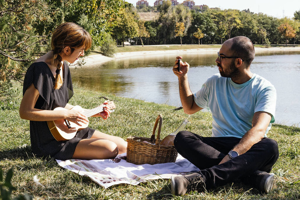 Pari istuu pöytäliinalla nauttien piknikistä, pelaa ukulelea ja ottaa kuvia - Valokuva, kuva