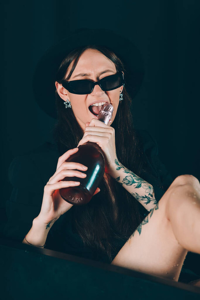 girl in a black jacket and hat posing in a black bath. A bottle of rose champagne wine in hand. Stylish sunglasses. Long naked sexy legs - Foto, afbeelding