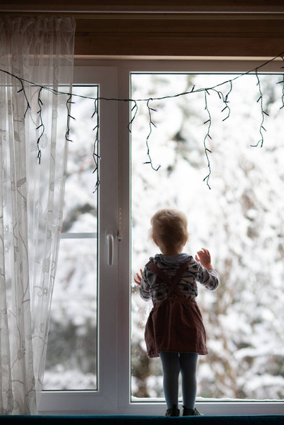 Παιδί κορίτσι στέκεται στο παράθυρο διακοσμημένο για τα Χριστούγεννα και βλέποντας το πρώτο χιόνι. Πρωτοχρονιάτικες διακοπές. Κάθετη μορφή - Φωτογραφία, εικόνα