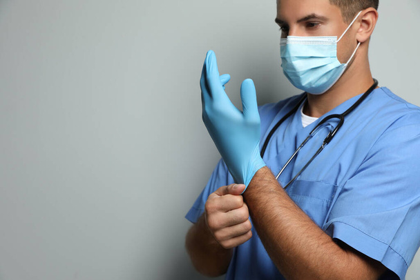 Doctor in protective mask putting on medical gloves against light grey background. Space for text - Fotografie, Obrázek