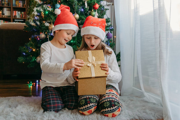 Egy fiú és egy lány karácsonyi kalapban kinyitnak egy ajándékdobozt egy karácsonyfa alatt. Ünneplés, ajándékok fogadása, gyerekek öröme. Csoda, jelen - Fotó, kép