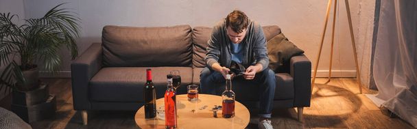 uomo ubriaco che tiene il portafoglio vuoto vicino al tavolo con bottiglie di bevande alcoliche a casa, banner - Foto, immagini