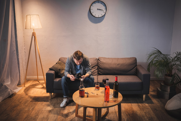 Зависимый от алкоголя мужчина держит пустой бумажник возле стола с бутылками дома - Фото, изображение