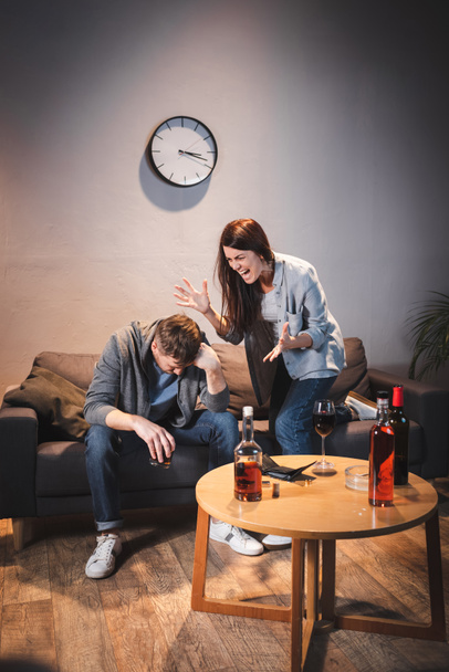 Θυμωμένη γυναίκα ουρλιάζοντας στο μεθυσμένο σύζυγο κοντά στο τραπέζι με ποτά αλκοόλ στο σπίτι - Φωτογραφία, εικόνα