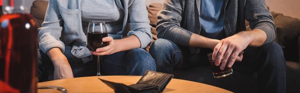 przycięty widok męża i żony siedzących z kieliszkami alkoholu w pobliżu pustego portfela, rozmyty pierwszy plan, baner - Zdjęcie, obraz
