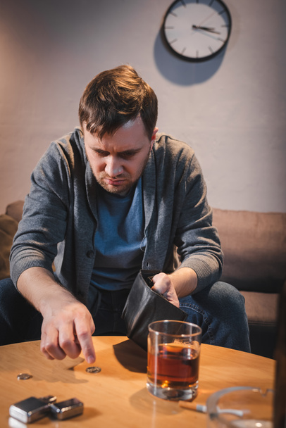 drunk man holding empty wallet near coins and glass of whiskey on blurred foreground - Photo, Image