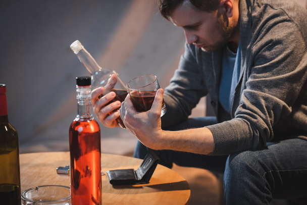 drunk man holding bottle and glass of whiskey near empty wallet on table - Zdjęcie, obraz