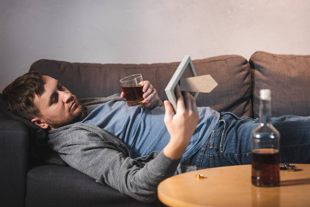 Aufgebrachter Mann liegt auf Sofa mit Fotorahmen und Glas Whiskey neben Flasche im verschwommenen Vordergrund - Foto, Bild