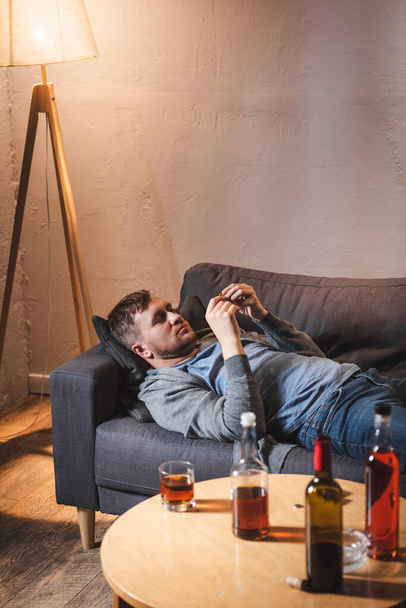 frustrado hombre acostado en el sofá cerca de botellas con bebidas alcohólicas en la mesa en casa - Foto, imagen