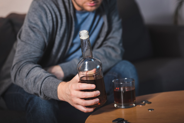 Teilaufnahme eines einsamen Mannes, der eine Flasche Whiskey in Glasnähe hält, verschwommener Hintergrund - Foto, Bild