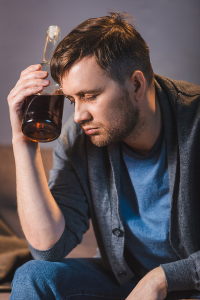 borracho, hombre deprimido sentado con botella de whisky cerca de la cabeza con los ojos cerrados - Foto, Imagen