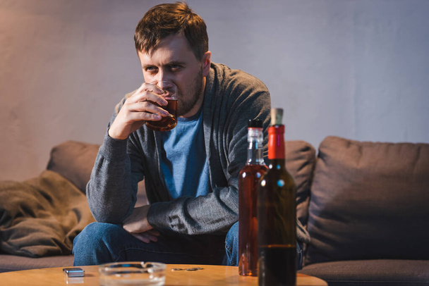 depressed, lonely man drinking whiskey near table with bottles of alcohol on blurred foreground - Foto, Imagem