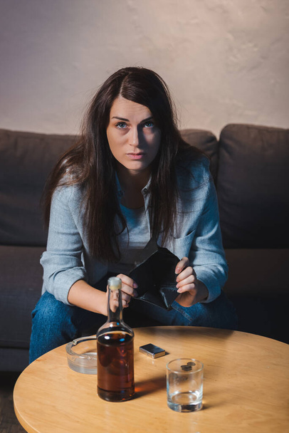 mujer deprimida mirando a la cámara mientras muestra la cartera vacía cerca de la botella de whisky en la mesa - Foto, imagen